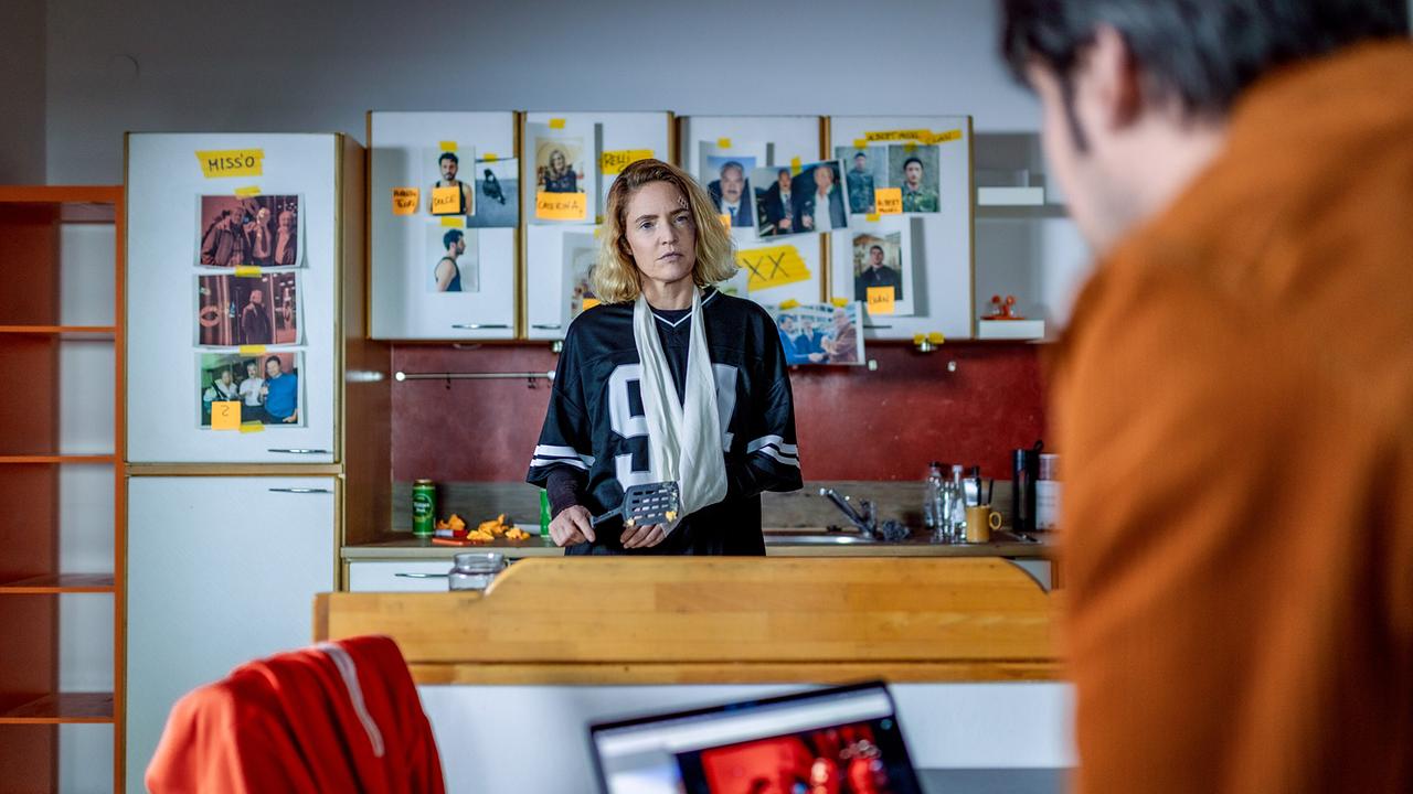 In einer Küche vor einer Wand voller Fotos und Notizen spricht Lisa Kuen mit jemandem.