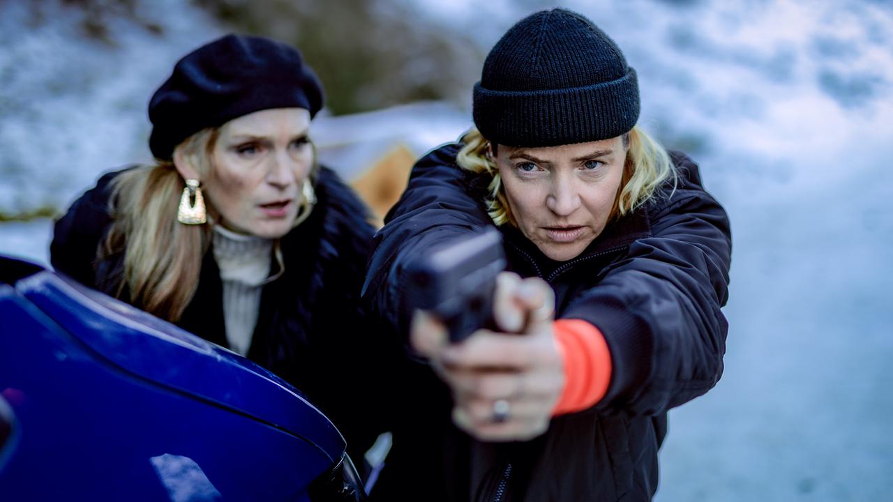Lisa Kuen richtet im Freien in einer schneebedeckten Landschaft eine Pistole auf jemanden, während Caterina Relli daneben steht.