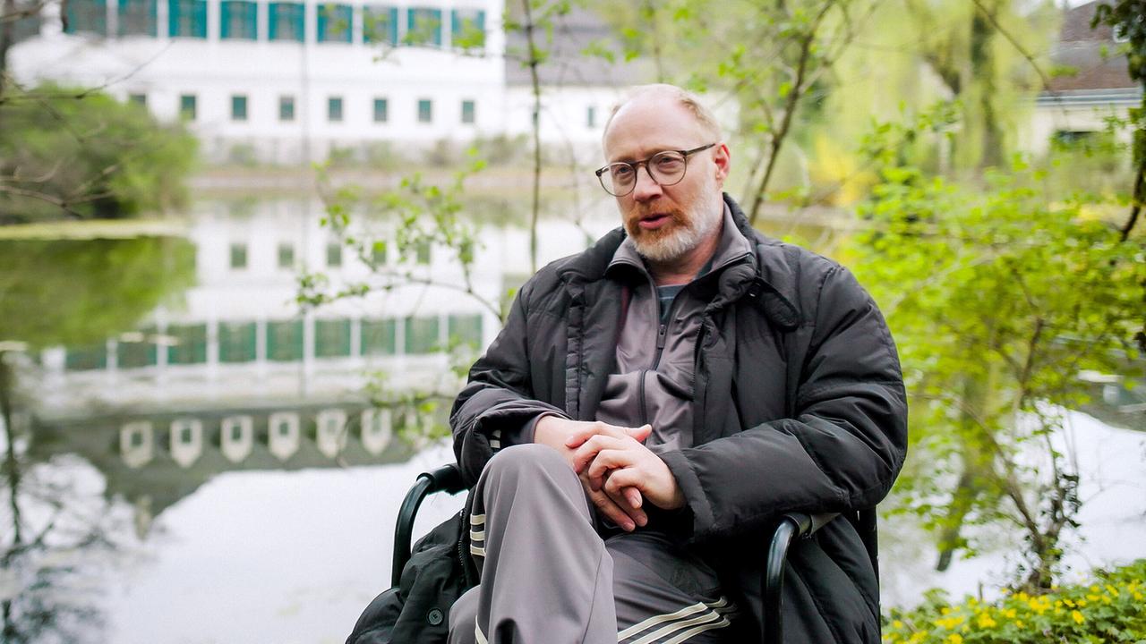 © ORF/AÖF Simon Schwarz im Interview sitzt in einem Sessel an einem Teich, im Hintergrund spiegelt sich ein großes helles Gebäude im Wasser.