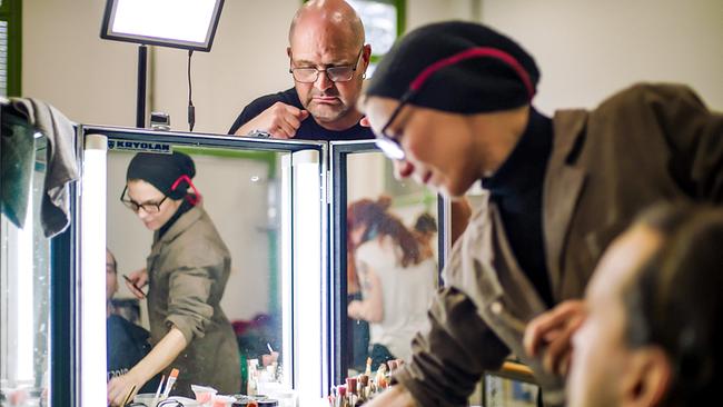 © ORF/AÖF Ein Mann wird bei einem Maskenworkshop vor beleuchteten Spiegeln von einer Maskenbildnerin vorbereitet, während im Hintergrund ein Mann zusieht.
