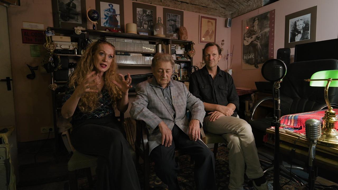 © ORF/AÖF Tizza Covi, Al Cook und Rainer Frimmel sitzen beim Interview in einem mit Blues-Memorabilien und Musikequipment vollgestellten Raum zum Film "Wiener Blues – The Loneliest Man in Town".
