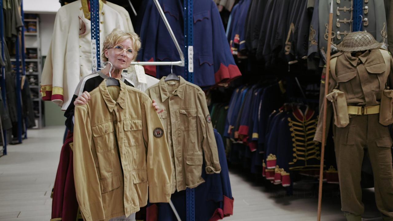 © ORF/AÖF Monika Buttinger zeigt in einem Fundus mehrere beigefarbene Uniformjacken aus dem Film "Bruno – Der junge Kreisky" vor Regalen mit historischen Kostümen.