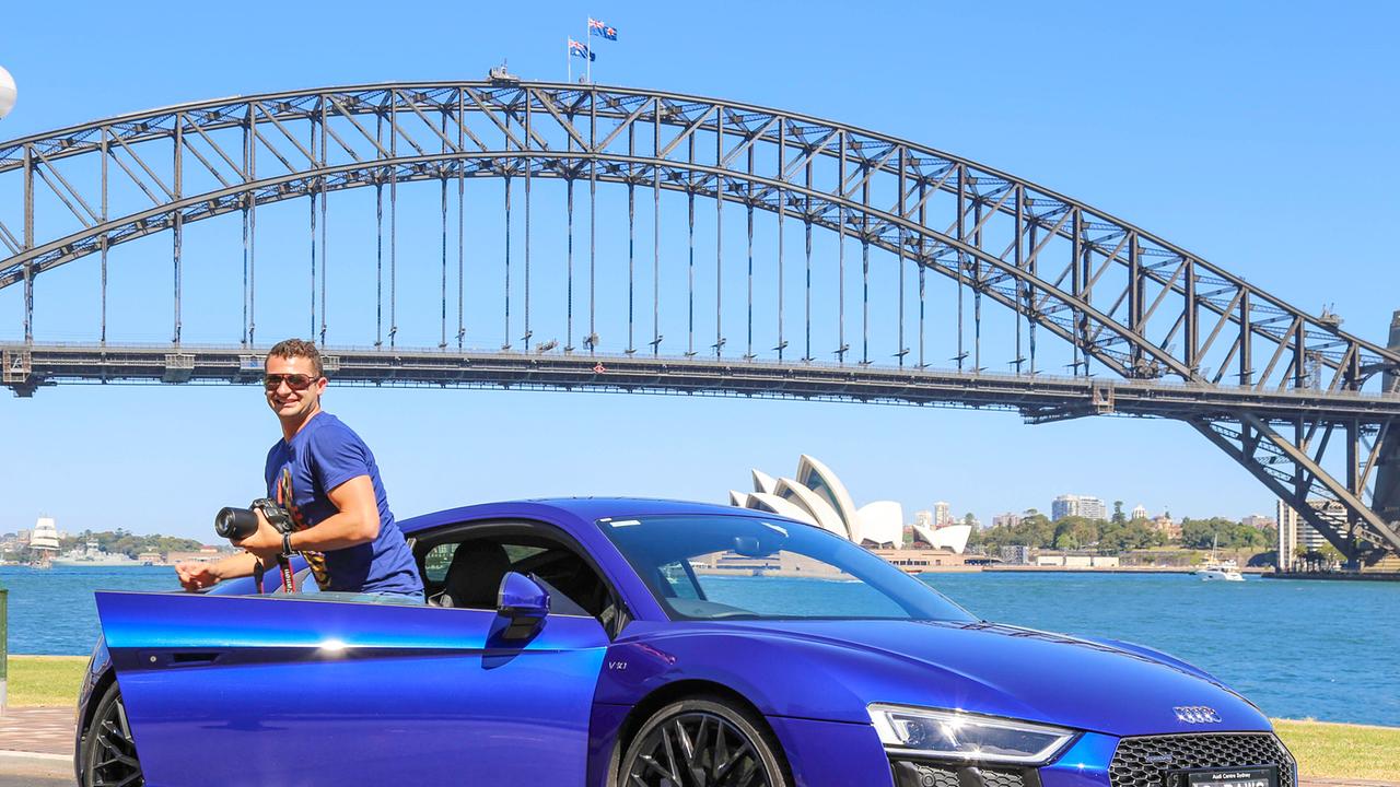 Anthony Koletti steigt von einem blauen Sportwagen aus, im Hintergrund Gewässer, eine hohe Brücke und das Opernhaus von Sydney