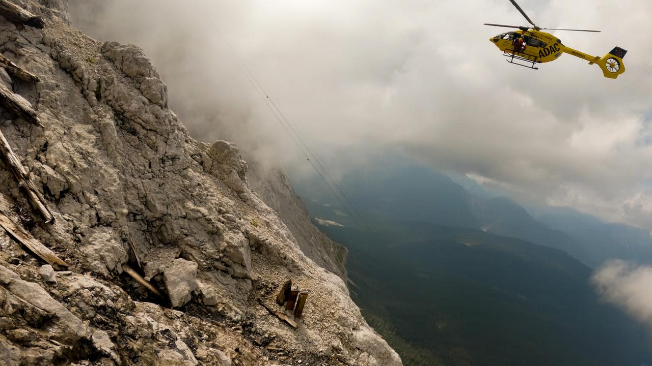 © ORF/BR/Timeline Production Ein Rettungshubschrauber schwebt bei einem Einsatz der Bergwacht Grainau über einer steilen, wolkenverhangenen Felswand in den Bergen.