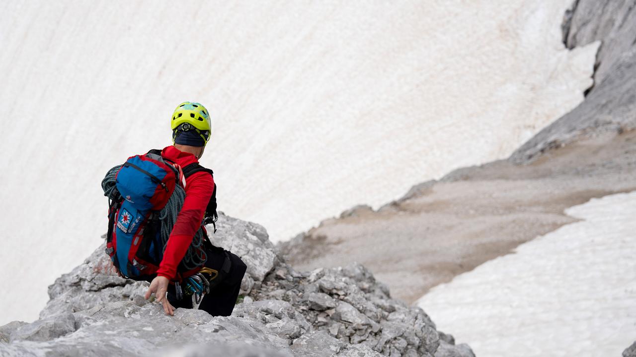 © ORF/BR/Timeline Production Ein Bergwachtler der Bergwacht Grainau bei einem Einsatz in der Nähe der Zugspitze sitzt in roter Jacke und gelbem Helm auf einem felsigen Vorsprung, mit Ausrüstung am Rücken, vor einer schneeweißen Bergwand.