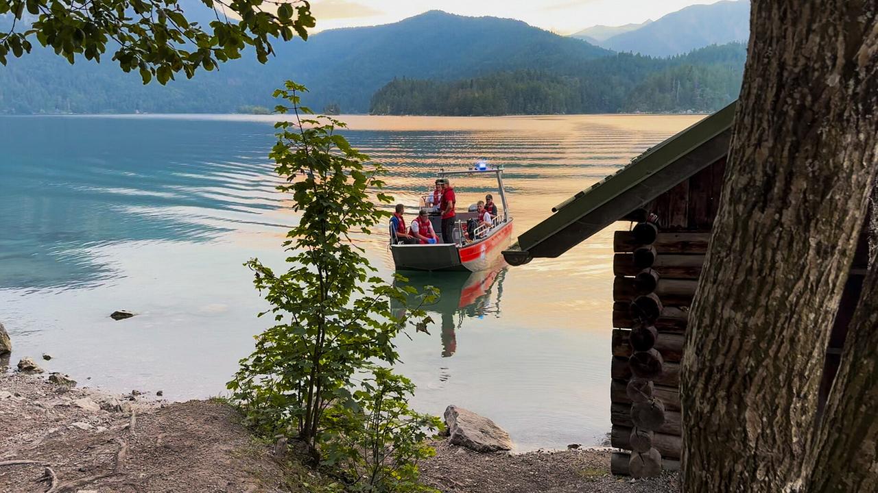 © ORF/BR/Timeline Production Die Bergwacht in roten Rettungswesten sitzt in einem Boot auf dem Eibsee, nahe einer kleinen Holzhütte am Ufer, vor einer Berglandschaft im Abendlicht.