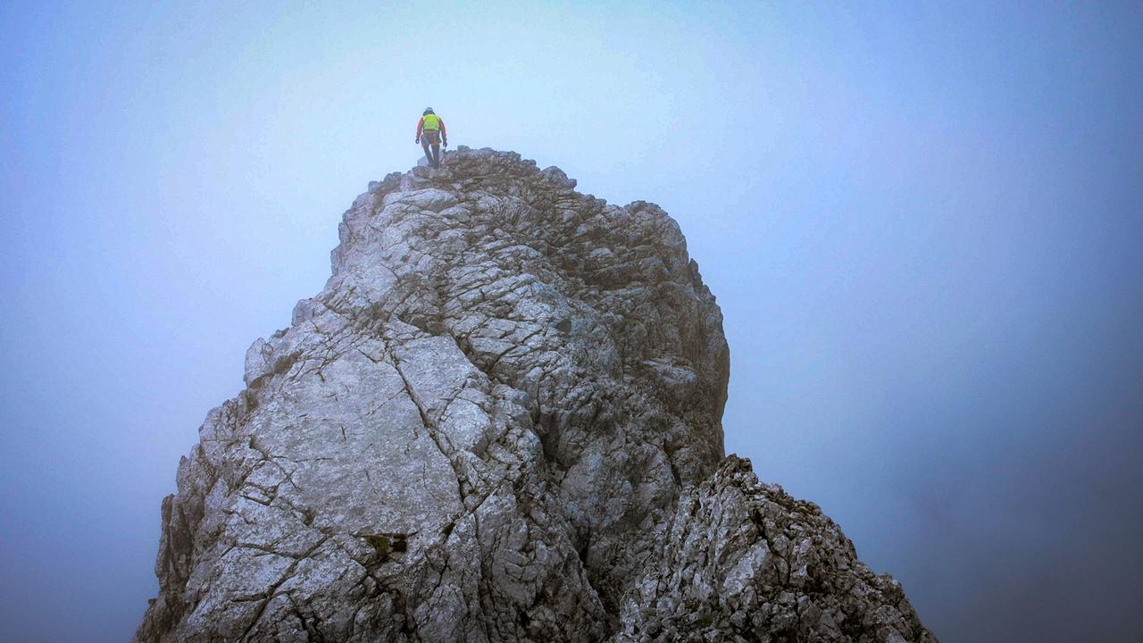 © ORF/BR/Timeline Production Einsatz der Bergwacht Ramsau am Watzmann: Eine einzelne Bergretter steht auf einem schroffen Felsgrat im Nebel, hoch über der Landschaft, umgeben von blau-grauer, wolkiger Atmosphäre.