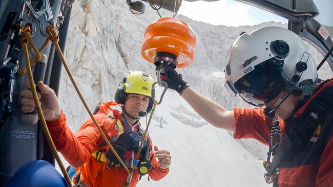 Andreas Pischl und ein weiterer Bergretter der Bergwacht Grainau in orangefarbenen Einsatzanzügen bereiten an einem Hubschrauber vor einer verschneiten Felswand eine Seilbergung vor.