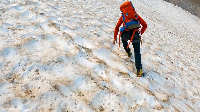 © ORF/BR/Timeline Production Ein Bergsteiger der Bergwacht Grainau mit orangefarbenem Rucksack steigt über ein steiles, teilweise verschmutztes Schneefeld.