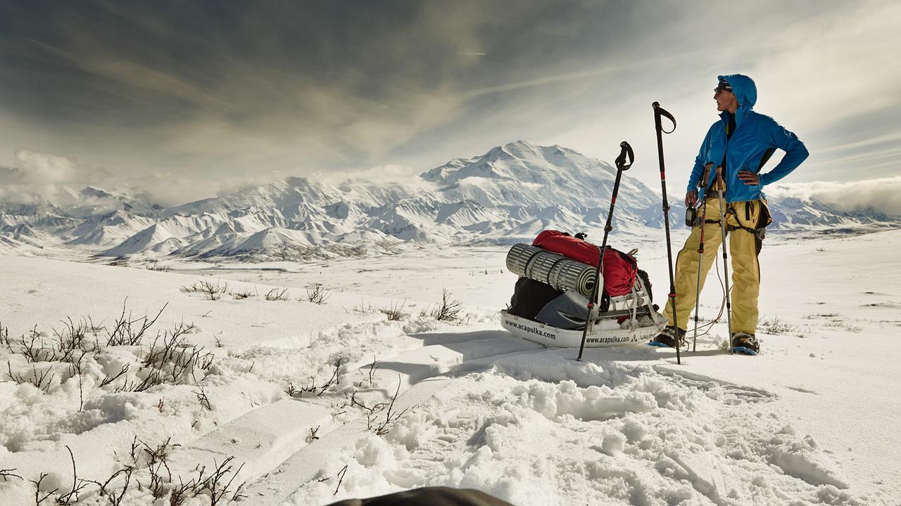 © ORF/ARD Plus/WDR Die Bergsteigerin Sylvia M. steht mit ihrer Ausrüstung auf einer verschneiten Ebene, im Hintergrund das Bergmassiv des Denali, des höchsten Bergs von Alaska