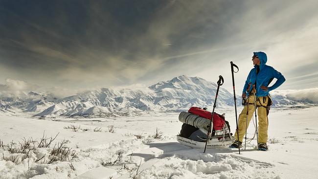 Die Bergsteigerin Sylvia M. steht mit ihrer Ausrüstung auf einer verschneiten Ebene, im Hintergrund das Bergmassiv des Denali, des höchsten Bergs von Alaska