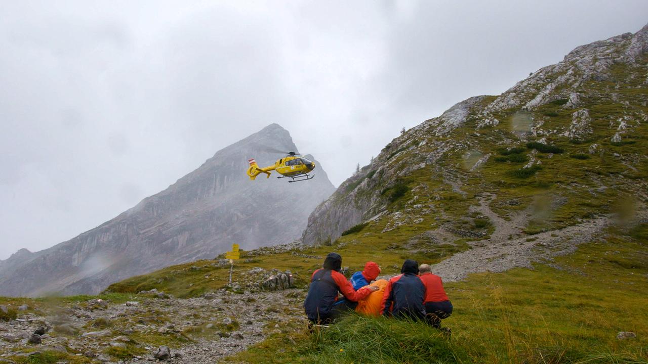 © ORF/BR/Timeline Production Einsatz der Bergwacht Ramsau: Ein gelber Rettungshubschrauber fliegt über nebelige Berglandschaft, während mehrere Personen in Regenjacken am Boden zusammengekauert sitzen.