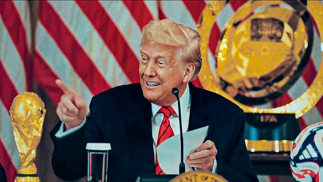 Donald Trump sitzt bei einer Pressekonforenz vor dem Mikrofon, deutet zu seiner rechten Seite und lächelt, auf dem Tisch vor ihm ein Pokal und ein Fußball