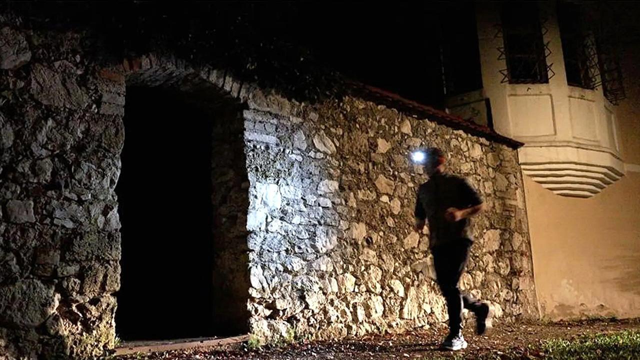 Nachgestellte Szene: In der Nacht läuft ein Mann mit Stirnlampe an einem Gebäuden mit alten Steinmauern vorbei, hinter ihm ein Erker des Hauses, vor ihm ein Tor in der Steinmauer