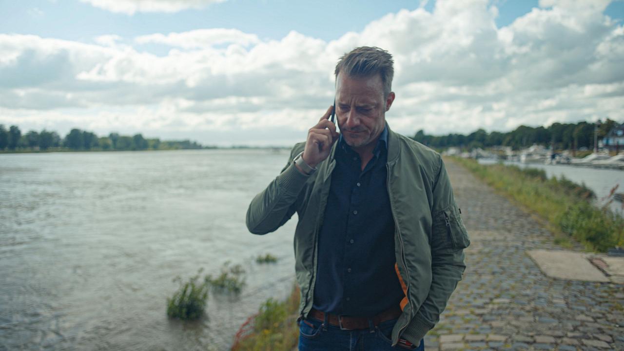 © ORF/One Gate/WDR/beetz brothers film production/Nikolaus von Schlebrügge Ein Mann in Jeans, blauem Hemd und grüner Jacke telefoniert am Handy, dabei steht am Ufer eines breiten Flusses, Boote sind in einem Seitenkanal zu sehen