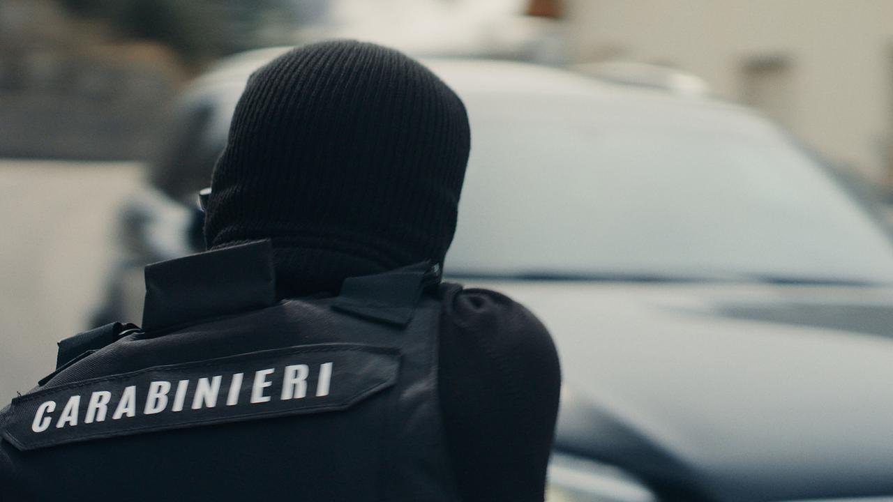 © ORF/One Gate/WDR/beetz brothers film production/Nikolaus von Schlebrügge Ein italienischer Polizist in Uniform, unter einer schwarzen Haube vermummt, hat ein Auto visiert, das unscharf im Hintergrund zu sehen ist
