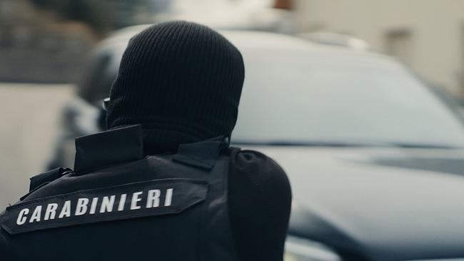 Ein italienischer Polizist in Uniform, unter einer schwarzen Haube vermummt, hat ein Auto visiert, das unscharf im Hintergrund zu sehen ist