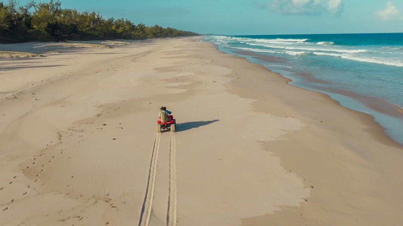 Zwei Personen fahren auf einem Quad einen paradiesischem Sandstrand entlang, links hinter den Dünen beginnt der Wald, rechts die Brandung des Meeres