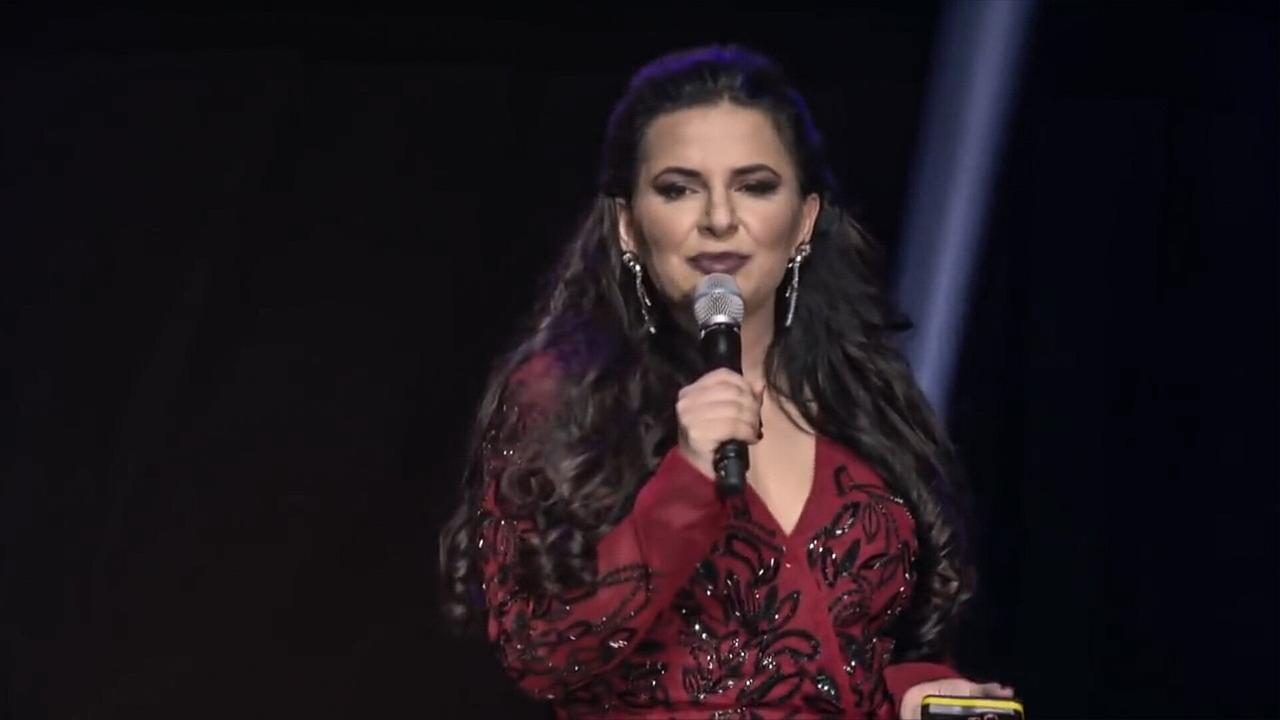 © ORF/a&o buero filmproduktion gmbh/WDR/Arte/Java Films/Martin Gasch Eine Frau mit langem dunkelbraunem Haar, in einem roten Kleid mit einem Mikrofon auf einer Bühne