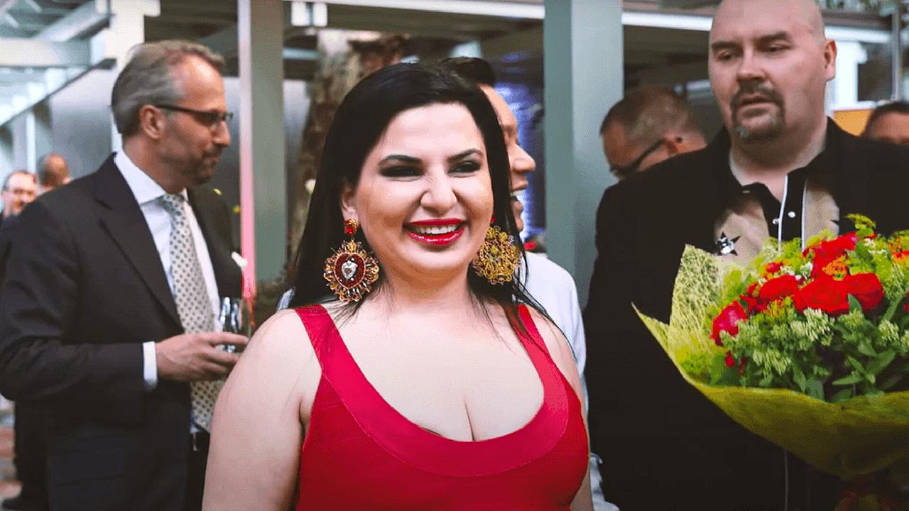 © ORF/a&o buero filmproduktion gmbh/WDR/Arte/Java Films/Till Vielrose Eine Frau mit schwarzen langen Haaren trägt ein rotes Kleid und großen Ohrschmuck, um sie stehen Männer mit Sektflöten in der Hand, einer hält einen Blumenstrauß