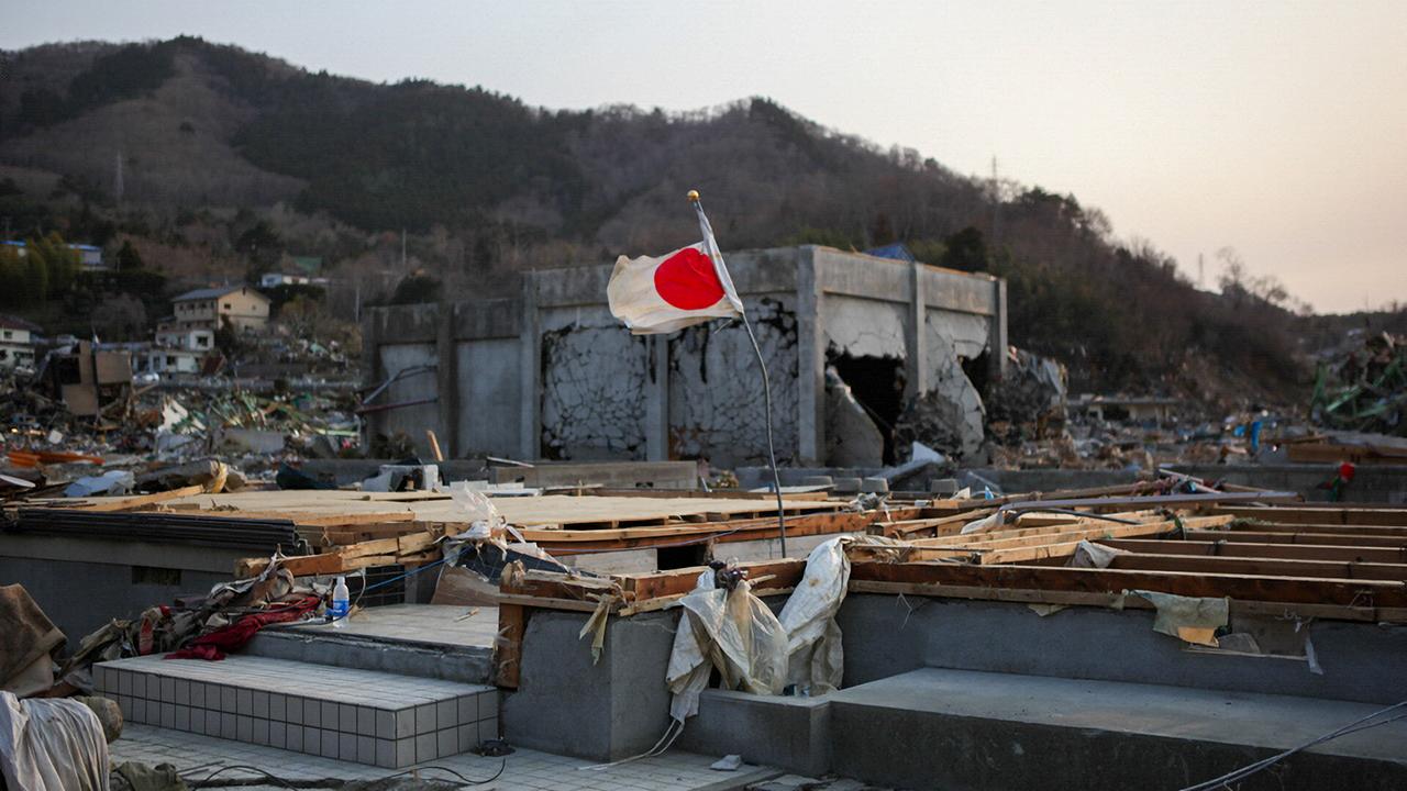 Von dem Tsunami zerstörtes Stadtgebiet, von einem Wohnhaus gibt es nur noch das Fundament, auf einem verbogenen Stab weht eine japanische Fahne 