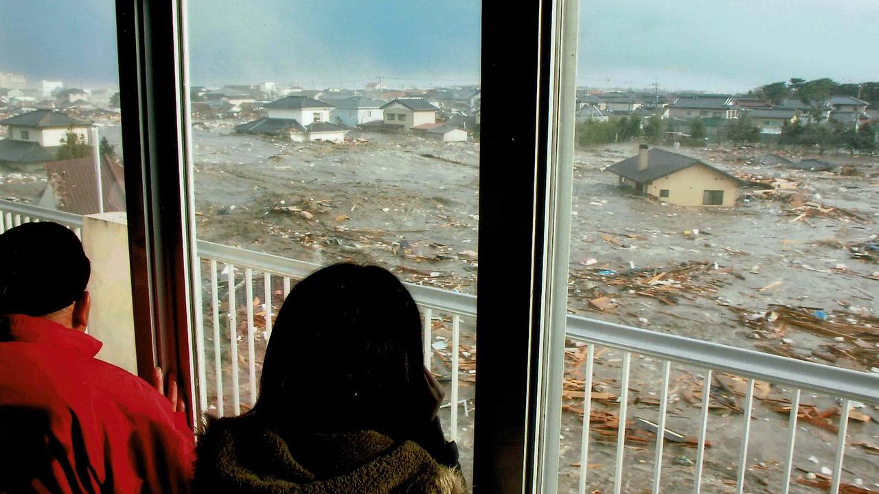Zwei Personen schauen durch die Fenster eines höher gelegenen Gebäudes auf den Tsunami, ein Wohnhaus und Unmengen von Treibgut werden vom Wasser weggespült
