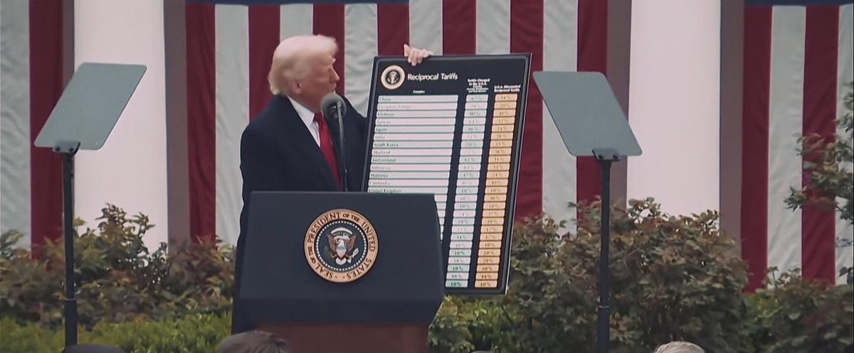 Donald Trump präsentiert er die Tafel mit Strafzöllen, vor ihm ein Rednerpult, hinter ihm die rot-weiße-Streifen der amerikanischen Flagge