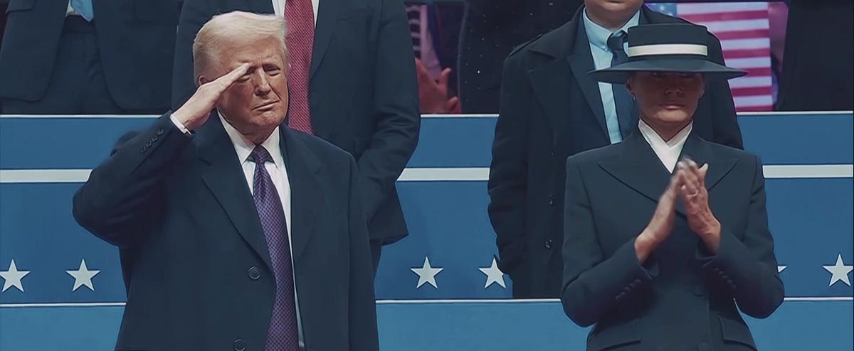 Bei einer Veranstaltung stehen Donald Trump und Melania Trump nebeneinander, er hält seine rechte Hand zum Salutieren an die Schläfe, sie applaudiert, beide blicken ins Publikum