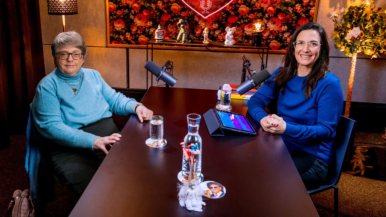 © ORF/Roman Zach-Kiesling Eine Studioaufnahme zeigt Dr. Helene Klaar und Nina Horowitz an einem dunklen Holztisch mit Mikrofonen vor einer dekorativen Wand mit Herzmotiv und der Aufschrift „Liebesgeschichten und Heiratssachen“.