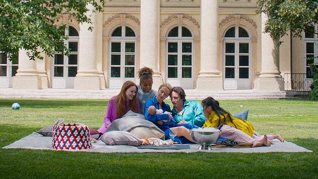 Familie Maynard entspannt auf einer Picknickdecke im Garten vor einem prächtigen, von Bäumen gesäumten Palais.