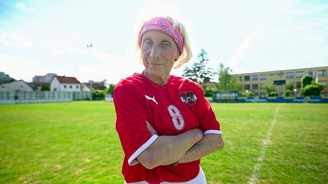 © ORF Christa Tauschek, eine ältere Frau im roten Fußballtrikot mit der Nummer 8, steht mit verschränkten Armen auf einem grünen Spielfeld unter blauem Himmel.