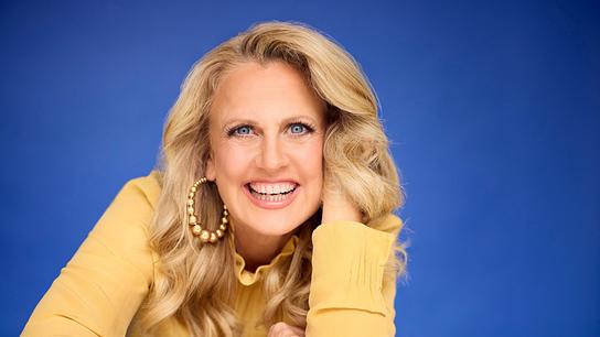 Porträtfoto von der lächelnden Barbara Schöneberger in gelber Bluse vor blauem Hintergrund 