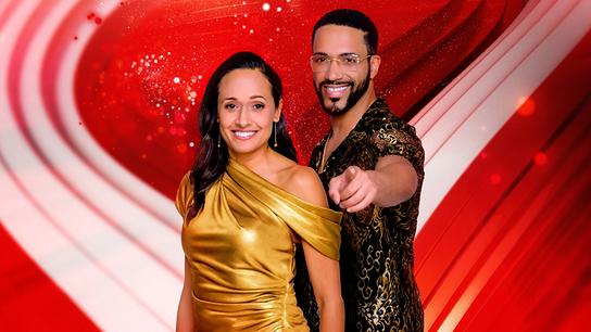 Alice Tumler und César Sampson in eleganter Kleidung stehen vor einem rot‑weiß gehaltenen Hintergrund.