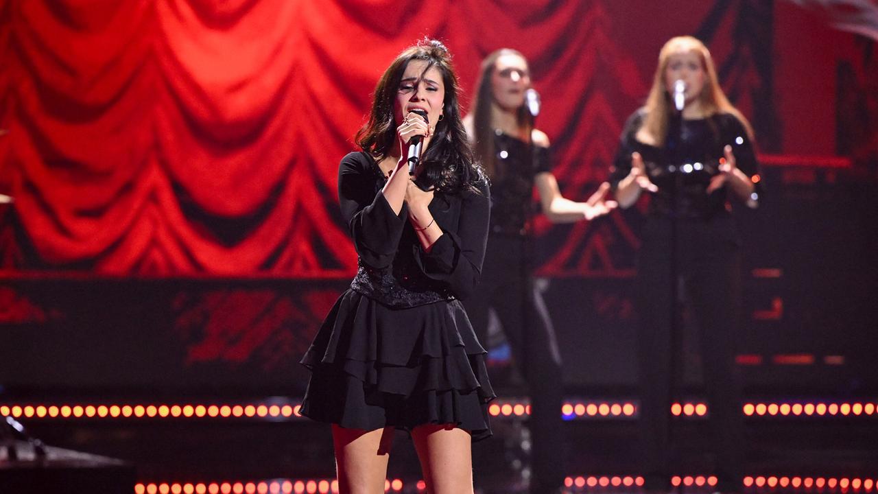 Vor rotem Samtvorhang singt Lena Schaur in schwarzem Outfit, während zwei Background-Sängerinnen dahinter stehen.