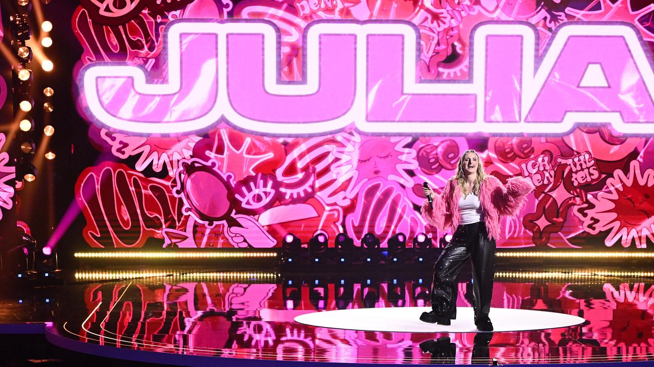 Vor einer knallpinken, graffitiartigen LED-Wand mit dem Schriftzug „JULIA“ performt Julia Steen im pinken Federjäckchen auf einem runden Podest.