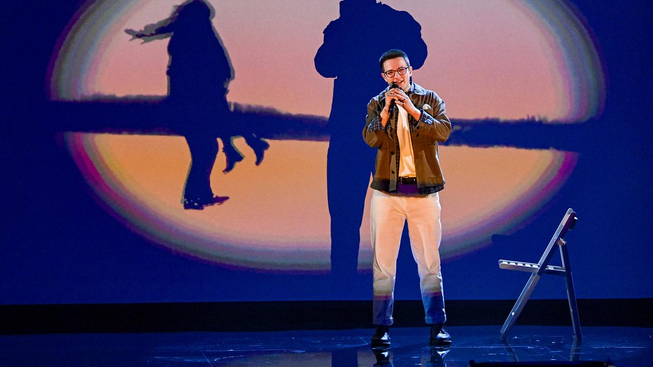 David Kurt singt vor einer Projektionsfläche mit großen Schattenbildern, daneben steht ein einzelner Stuhl auf der Bühne.