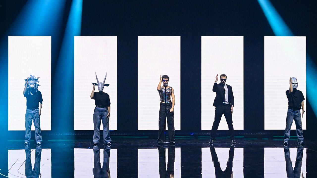 Fünf Personen von Cosmó stehen in einer Reihe vor hohen, weißen Lichtpaneelen, einige halten Masken oder Requisiten in die Höhe.