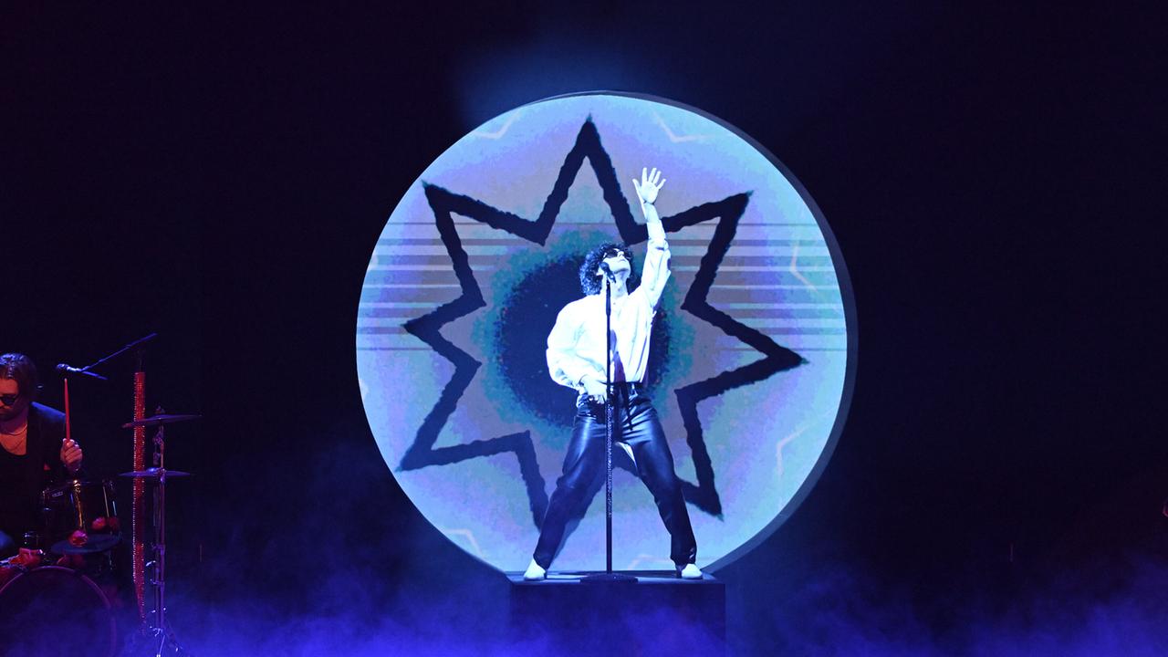 Nikotin steht auf einem Podest vor einem kreisrunden, sternförmigen Hintergrund und streckt eine Hand nach oben.