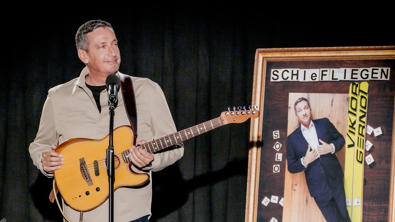 © ORF/televisionair Viktor Gernot steht mit einer Gitarre auf einer Bühne neben einem großen Plakat, das eine humorvolle Ski‑Motiv-Gestaltung für sein Solo‑Programm "Schiefliegen" zeigt.