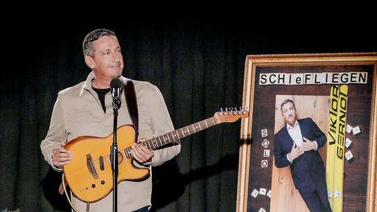 Viktor Gernot steht mit einer Gitarre auf einer Bühne neben einem großen Plakat, das eine humorvolle Ski‑Motiv-Gestaltung für sein Solo‑Programm "Schiefliegen" zeigt.