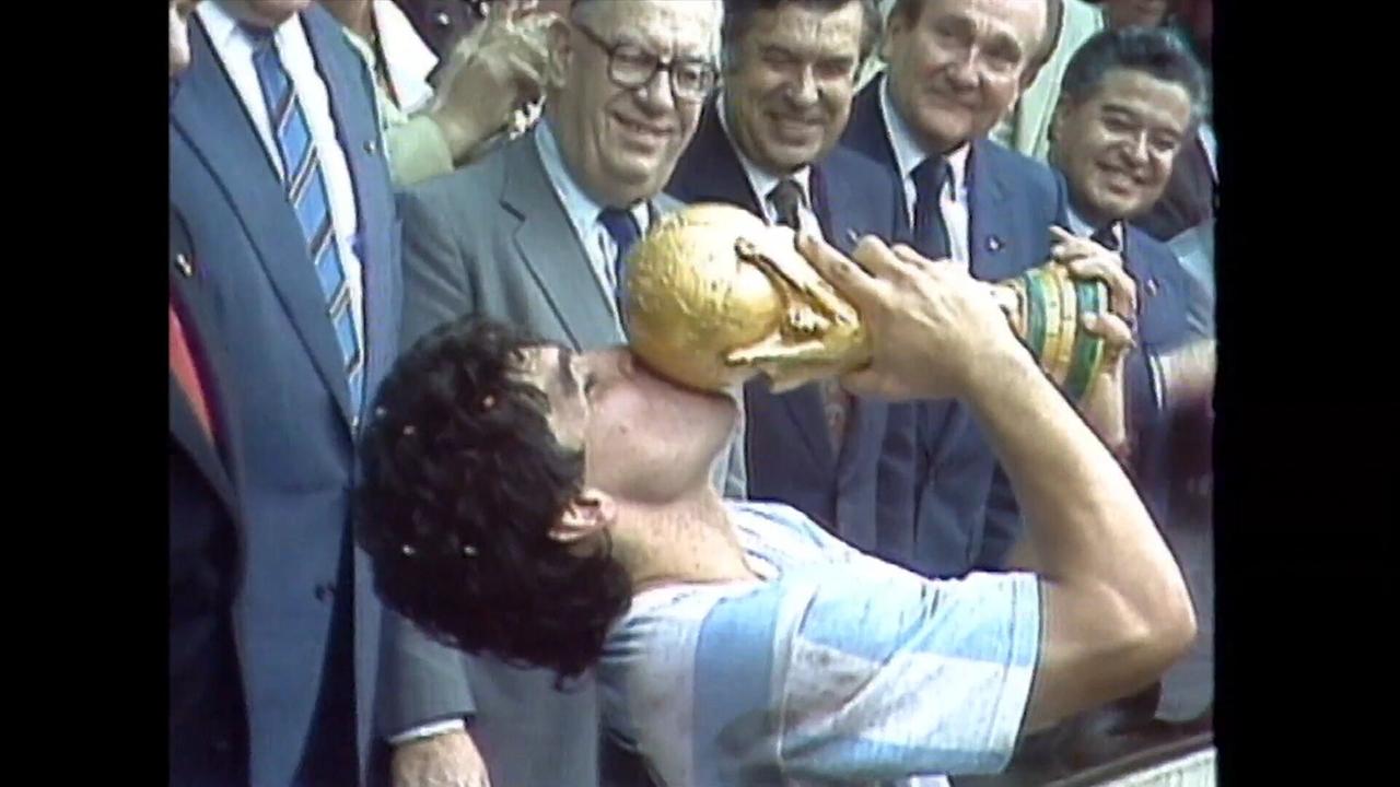 Diego Maradona im Trikot küsst den WM-Pokal 1986 in Mexiko, umgeben von Anzugträgern.