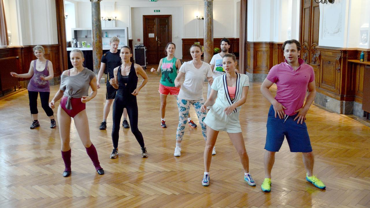 © ORF/MR Film/Hubert Mican Eine Gruppe von Menschen, unter ihnen Nicoletta, Waltraud, Maria, Caroline und Marko, trainiert gemeinsam in einem großen Saal und führt Fitnessbewegungen aus.