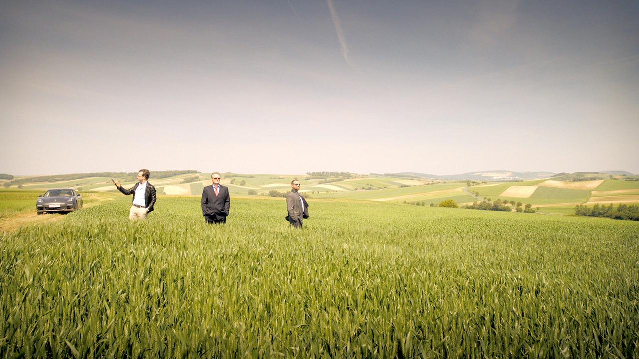 © ORF/MR Film Bertram Selig, Hadrian Melzer und Georg Schneider stehen in einem grünen Feld vor einer sanften Hügellandschaft, während ein Auto am Rand geparkt ist.