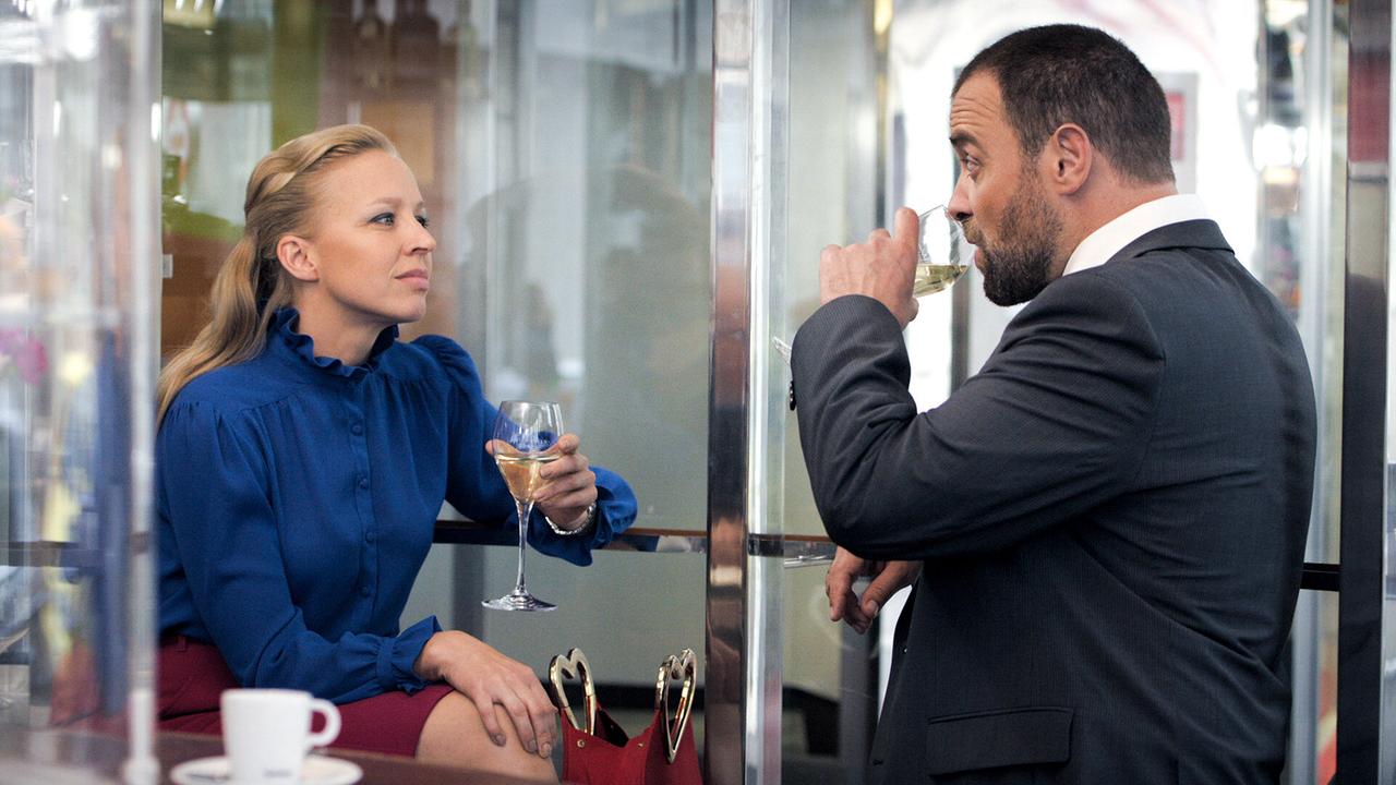 Nicoletta Huber in blauer Bluse und Georg Schneider im Anzug trinken in einer Bar Wein und unterhalten sich.