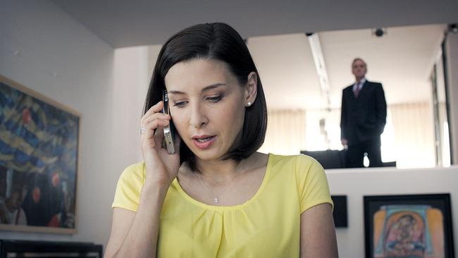 © ORF/MR Film Caroline Melzer in gelbem Oberteil telefoniert am Handy in einem Wohnzimmer, während im Hintergrund Hadrian Melzer im Anzug auf der oberen Wohnebene steht.