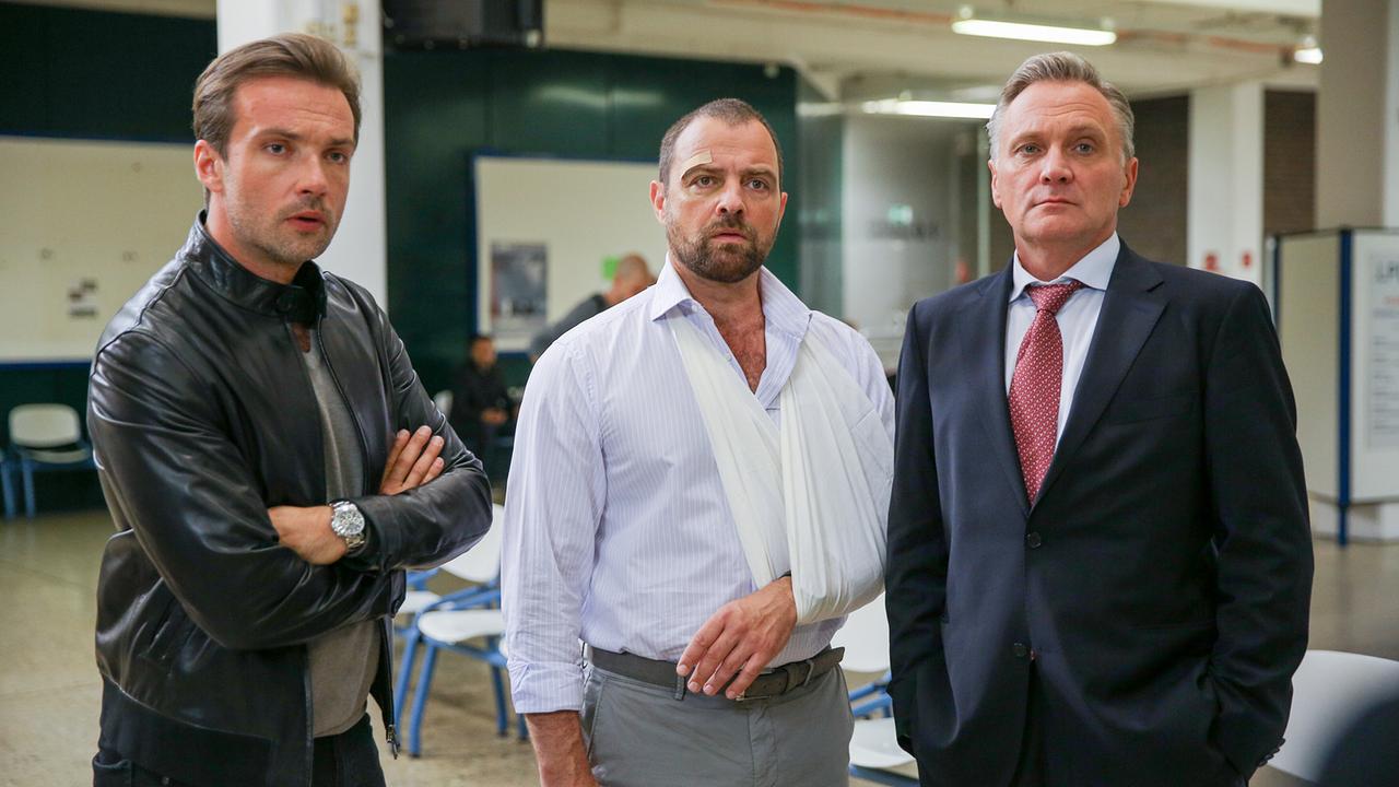 Bertram Selig, Georg Schneider mit einem Arm in der Schlinge und Hadrian Melzer im Anzug stehen in einem hellen Innenraum und schauen angespannt nach vorne.