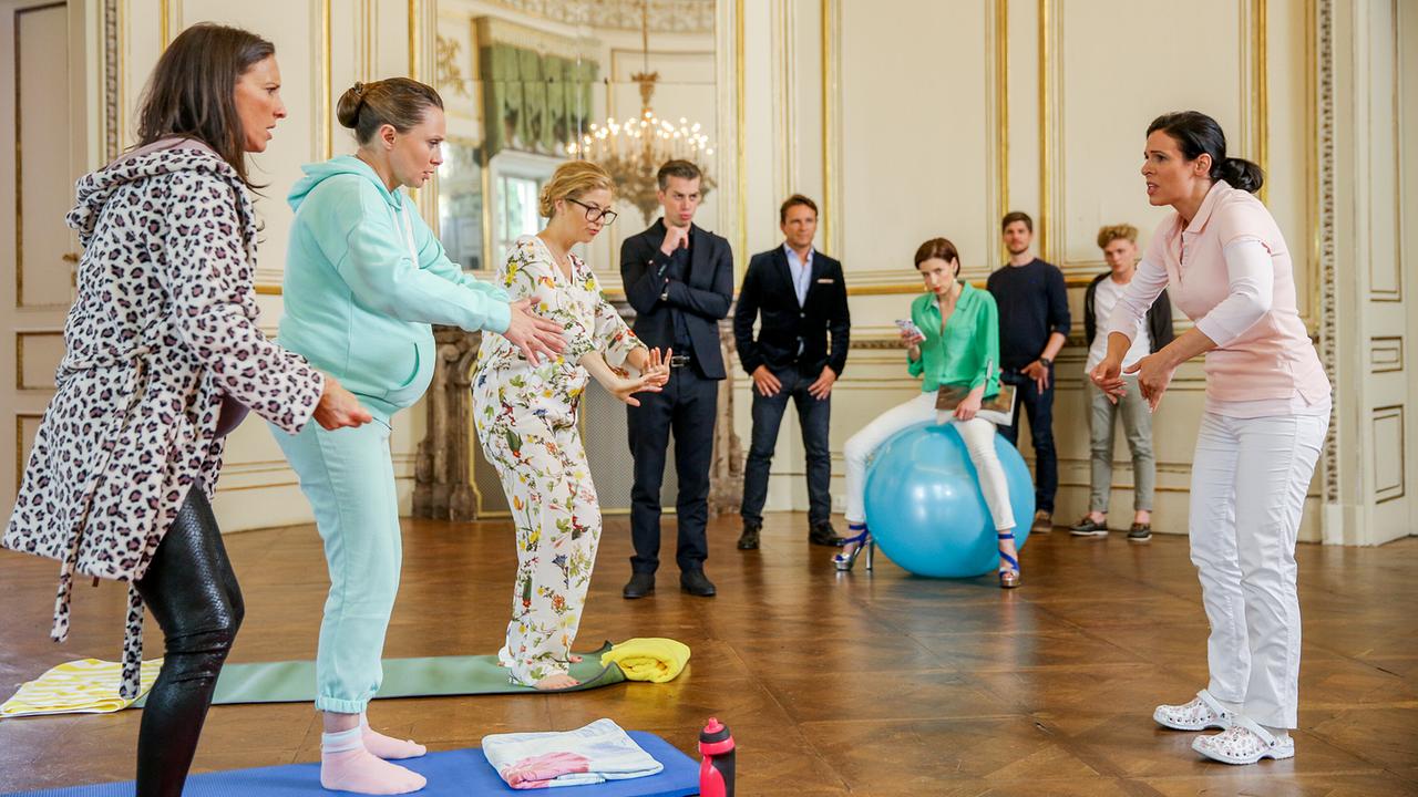 Waltraud Steinberg, Maria Schneider und Vanessa beim Schwangerschaftsturnen in einem eleganten Raum. An der Wand schauen Günter, Caroline Melzer auf einem Gymnastikball, Simon Schneider und zwei weitere Personen zu.