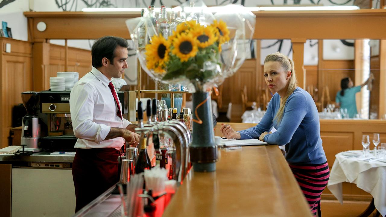 Nicoletta steht an einer Restaurantbar und blickt nachdenklich auf ein großen Sonnenblumenstrauß im Vordergrund, der auf der Theke steht. Antonio steht hinter der Bar.