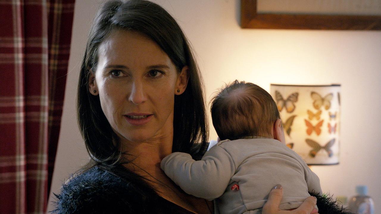 © ORF/MR Film Nahaufnahme von Waltraud, die in einem Wohnraum ein Baby auf dem Arm trägt.