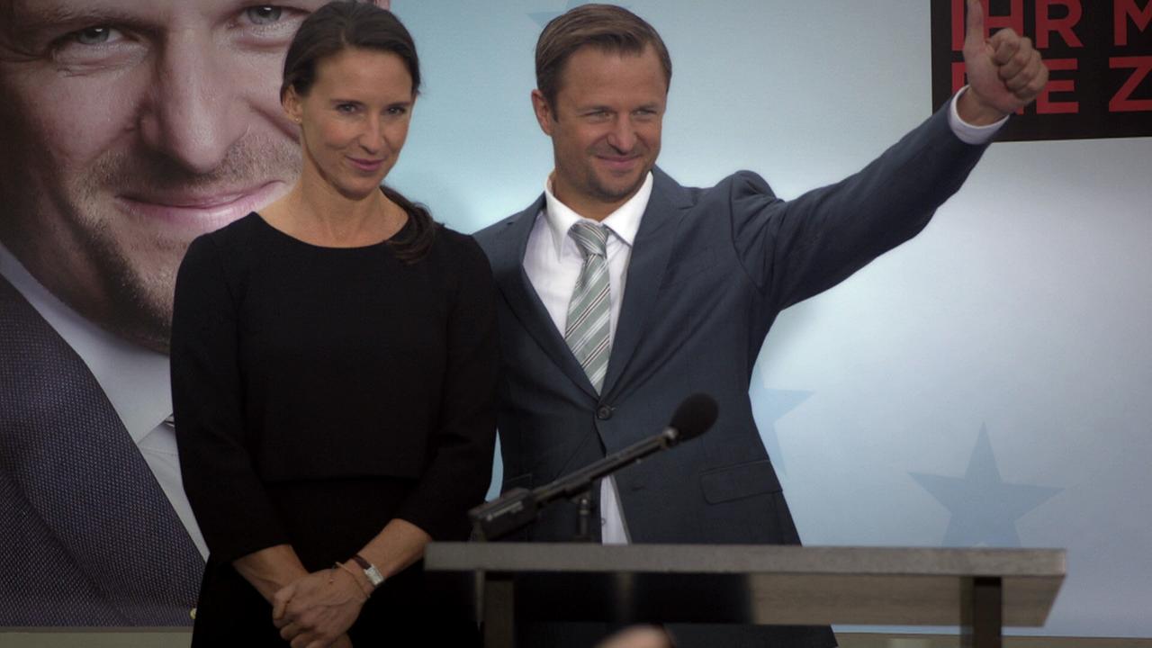 © ORF/MR Film Waltraud Steinberg steht vor einem Wahlplakat neben Joachim Schnitzler am Rednerpult, der mit erhobenem Daumen posiert.